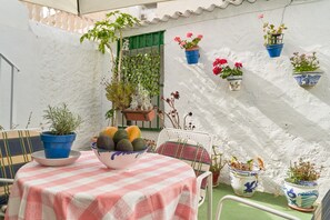 A table with a checkered tablecloth, potted plants, and a bowl of fruit on a balcony with white walls and a green door.