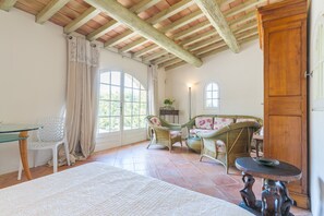 A room with a high wooden ceiling, a large window with curtains, wicker furniture, a round table, and a wooden door.