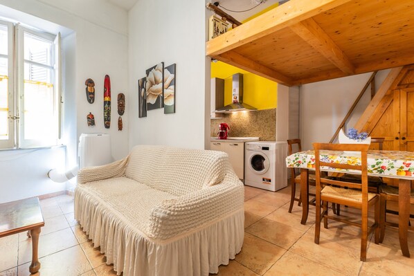 A cozy room with a sofa, a washing machine, a dining table with chairs, and a window with curtains. - Apartment 'Casa Musio' (Cagliari)