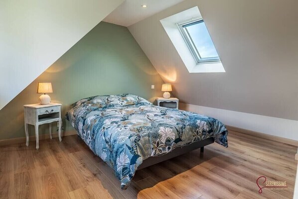 Une chambre avec un plafond en pente, un puits de lumière, un lit recouvert d’une couvre-lit à motifs floraux, deux tables de chevet avec des lampes et un plancher en bois.