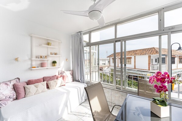 A hotel room with a bed, a chair, a table, a window with a view of buildings, and a ceiling fan.