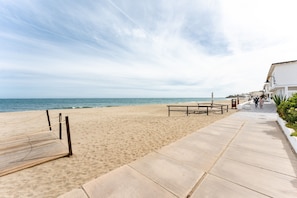 Ein Strand mit einer Holzterrasse, einem Betonweg und Gebäuden im Hintergrund.