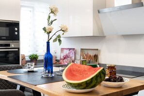 Una cocina con una mesa, un jarrón con flores, una tajada de sandía y un tazón de cerezas.