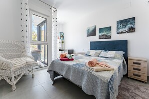 Un dormitorio con cama, una silla de mimbre, una mesita de noche y una lámpara, además de una obra de arte en la pared.