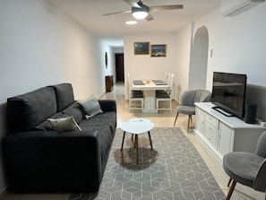 A living room with a sofa, a TV, a dining table with chairs, and a ceiling fan. - Apartment 'Nuevo Acapulco' with Mountain View, Shared Pool and Shared Garden (Fuengirola)