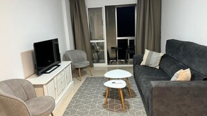 Ein modernes Wohnzimmer mit Flachbildfernseher, grauen Sesseln, einem grauen Sofa, einem kleinen weißen Tisch und einem Deckenventilator.