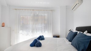Une chambre d’hôtel avec un lit, une fenêtre avec des rideaux légers, une table de chevet et un climatiseur fixé au mur.