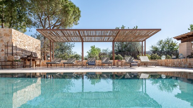 Una piscina de aguas cristalinas, una pérgola de madera, sillas de descanso y muros de piedra.