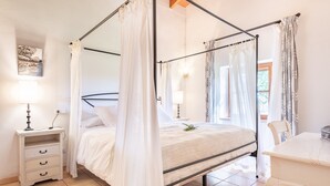 Una cama con dosel, una mesita de noche con lámpara, un pequeño aparador y vistas al exterior a través de una ventana.