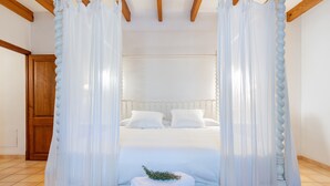 Une chambre à coucher avec un lit, des rideaux blancs, une porte en bois et une petite table sur laquelle se trouvent une serviette et quelques plantes.
