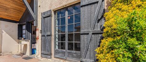 A building with a rustic exterior, large windows with shutters, a wooden ceiling, and a small outdoor area with a table and plants.
