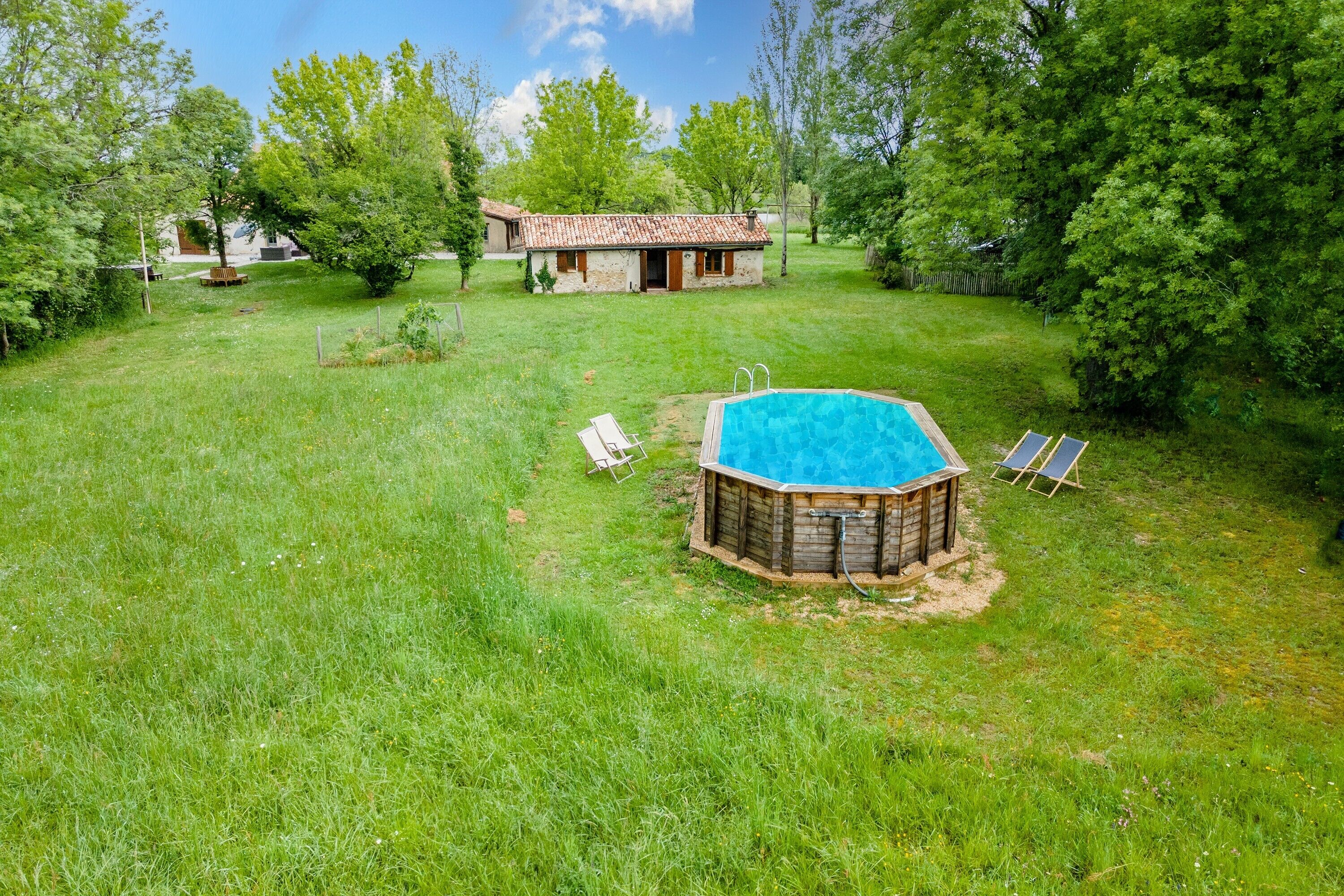 A wooden hot tub with a blue pool, surrounded by a grassy yard with two lounge chairs and a small building in the background.