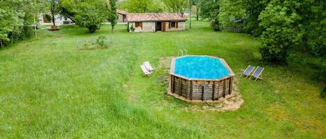A wooden hot tub with a blue pool, surrounded by a grassy yard with two lounge chairs and a small building in the background.