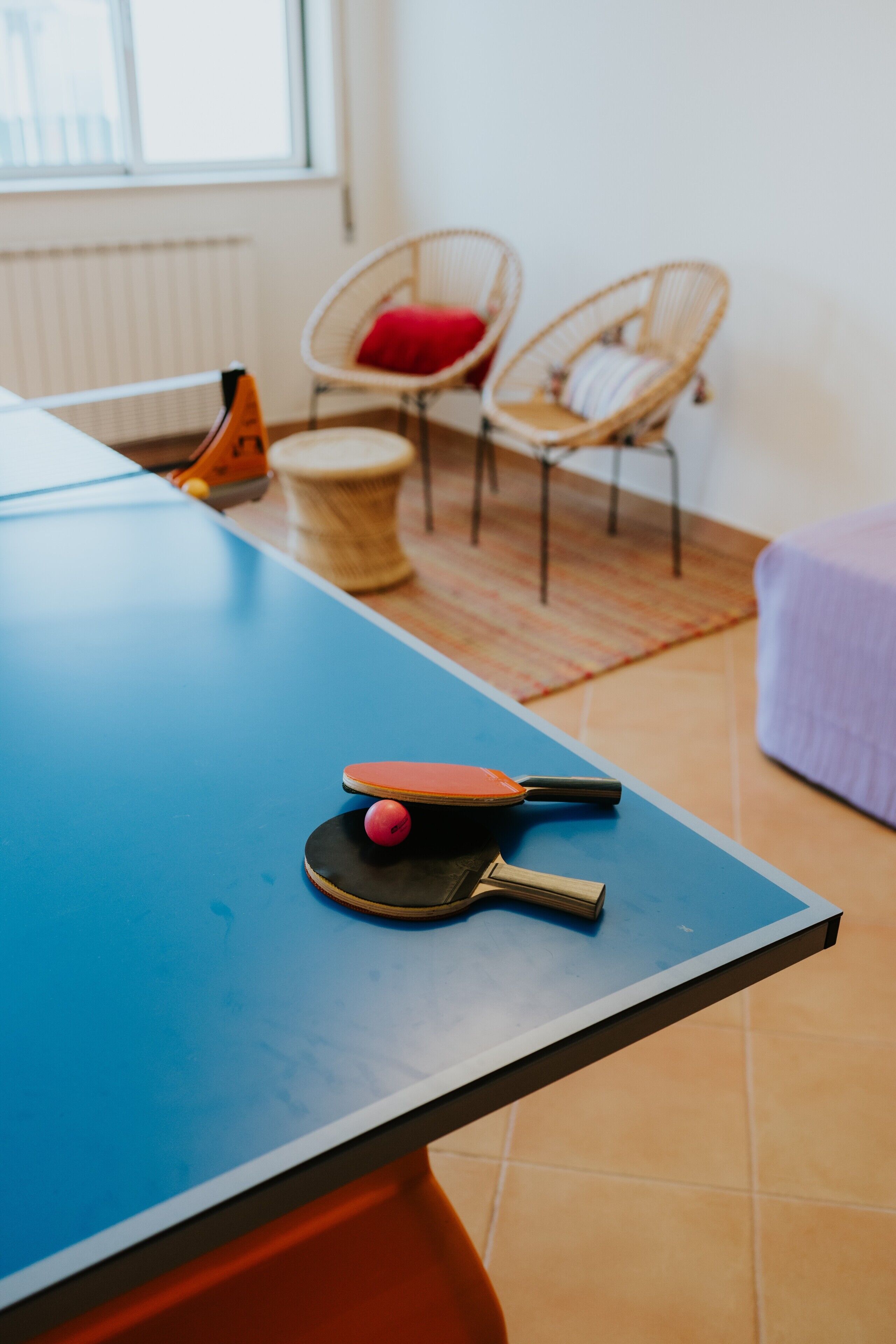 A ping pong table with paddles and a ball, wicker chairs with cushions, a small drum, and a bed with a purple blanket.