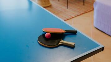A ping pong table with paddles and a ball, wicker chairs with cushions, a small drum, and a bed with a purple blanket.
