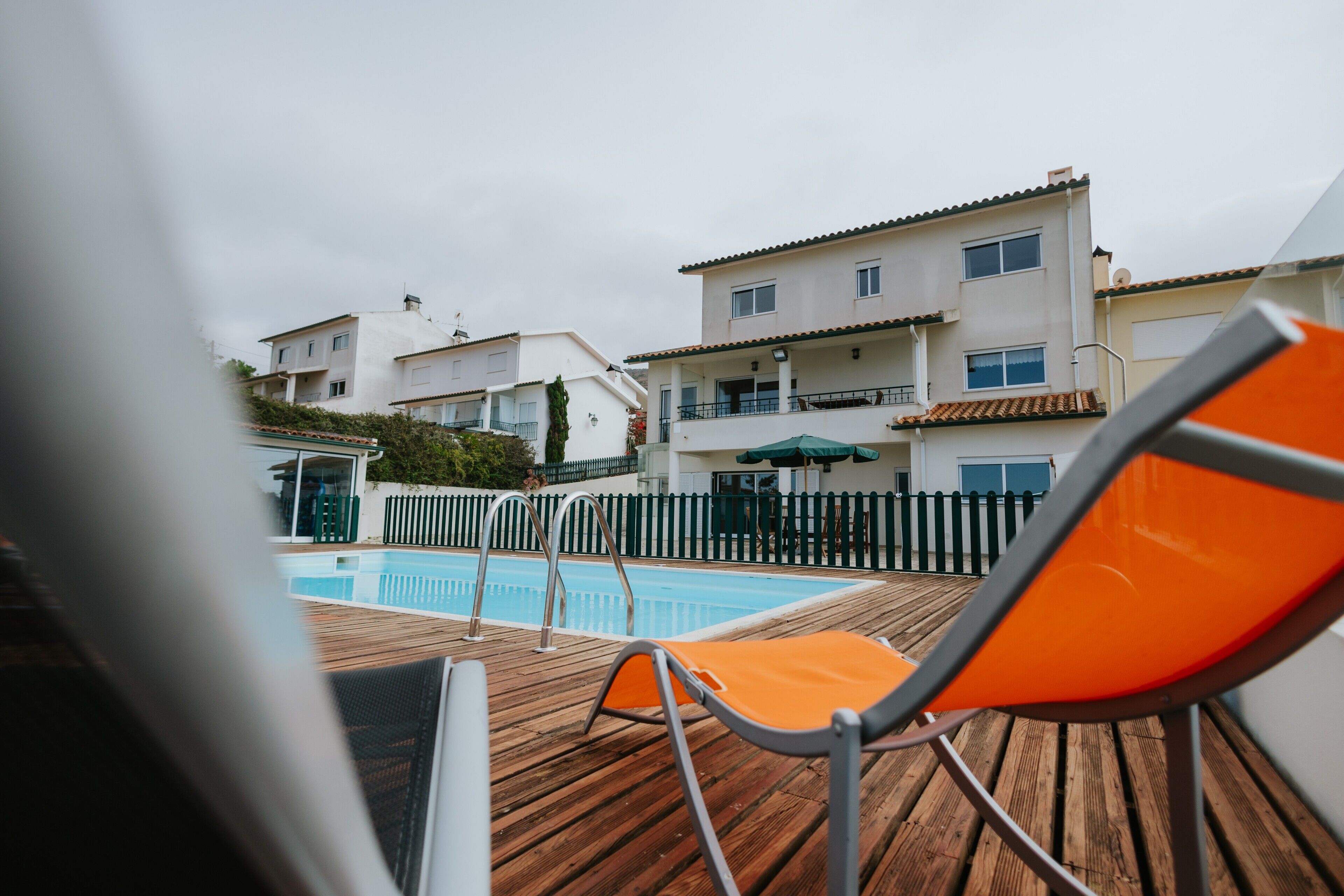 Una terraza de madera con piscina, una silla y un edificio con balcones al fondo.