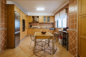 A kitchen with wooden cabinets, a stainless steel refrigerator, a dining table with chairs, and a window with curtains. - Holiday Home 'Casa Das Camarinhas' with Sea View, Private Pool and Wi-Fi (Murtinheira)