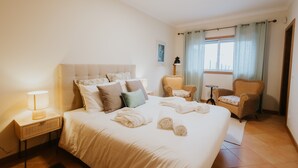Een hotelkamer met een groot bed, een nachtkastje met een lamp, twee rieten stoelen, een kleine tafel en een raam met gordijnen.