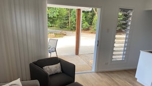 A living room with a sofa, an ottoman, and a chair with a cushion. There is a sliding glass door leading to an outdoor area with greenery. - Fantastic views, comfortable beds. Close to Banya Bathhouse Spa & Crystal Castle (Mullumbimby)
