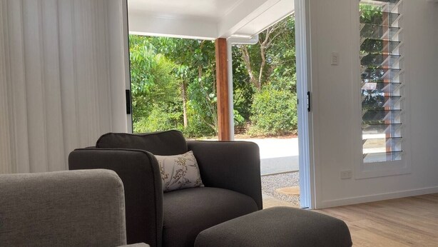 A modern living room with a sofa and ottoman, a sliding glass door leading to a garden, and a window with blinds. - Fantastic views, comfortable beds. Close to Banya Bathhouse Spa & Crystal Castle (Mullumbimby)