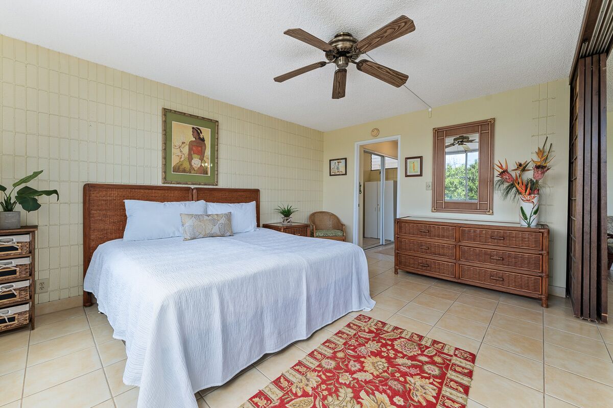 A bedroom with a large bed, a wooden headboard, a ceiling fan, a red patterned rug, a dresser, and a mirror.