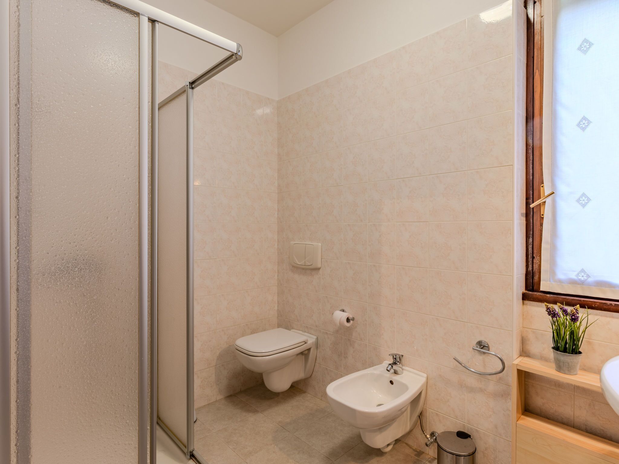 A bathroom with a glass shower enclosure, a toilet, a bidet, and a sink with a wooden shelf.