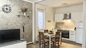 Une cuisine moderne avec des armoires blanches, un four encastré et un évier. On y trouve une table à manger avec quatre chaises et un panier de fruits. Un téléviseur à écran plat est posé sur une armoire blanche, à gauche.