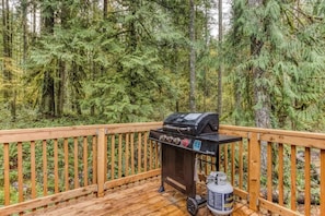 A deck with a grill and propane tank, surrounded by dense forest.