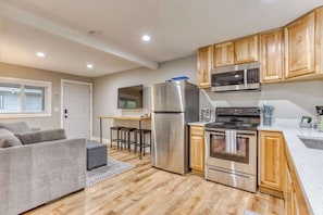 A modern kitchen with stainless steel appliances, wooden cabinets, and a grey sofa in a living area.