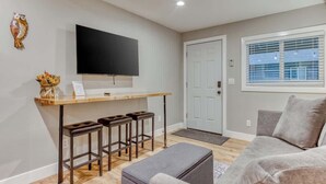 Una sala moderna con una televisión de pantalla plana, un escritorio de madera con taburetes, un sofá gris y una puerta blanca.