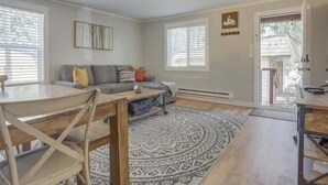 Un salon avec un canapé gris, une table basse et un tapis à motifs. On y trouve également une table à manger avec des chaises, un luminaire au plafond et une porte donnant sur l’extérieur.