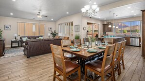 A spacious living room with a brown leather sofa, wooden dining table with chairs, and a kitchen area with a large window.