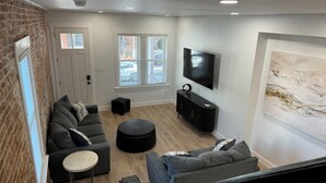 A modern living room with a brick wall, a sofa set, a black ottoman, a TV, and a large abstract painting.