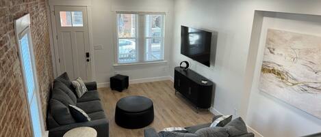 A modern living room with a brick wall, a sofa set, a black ottoman, a TV, and a large abstract painting.