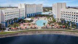 A hotel with a large central pool surrounded by palm trees and umbrellas, multiple buildings with a modern design, and a clear view of the surrounding landscape.