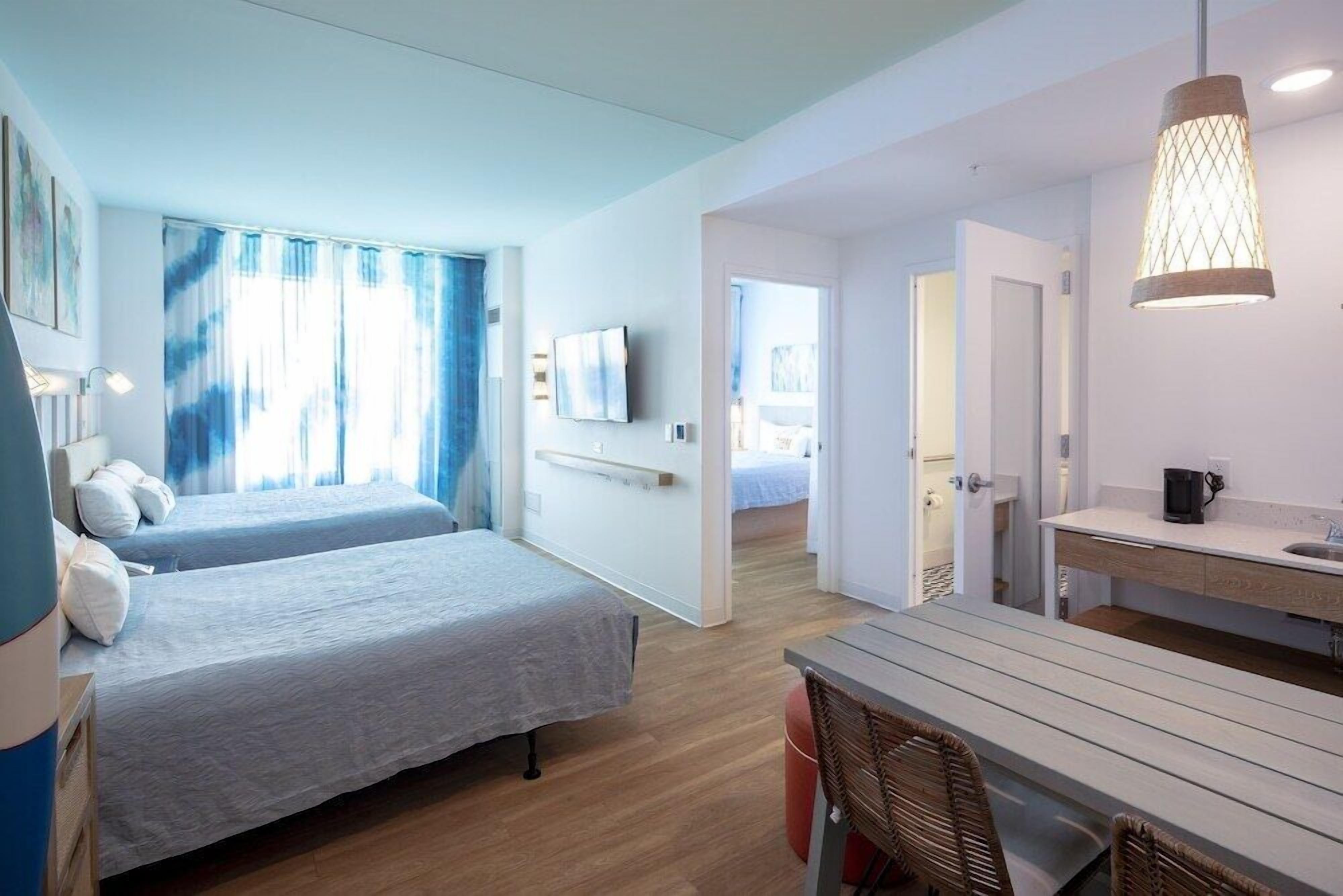 A modern hotel room with two beds, a dining area, and a kitchenette.