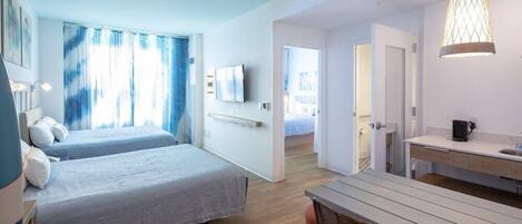 A modern hotel room with two beds, a dining area, and a kitchenette.