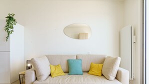 Un salon aux couleurs neutres, avec un canapé beige, deux coussins jaunes et un coussin turquoise, une petite table d’appoint, un lampadaire, un miroir et une plante.