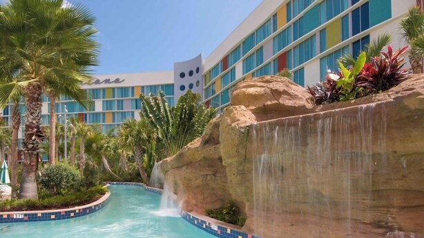 Een hotelzwembad met een waterval, omringd door palmbomen en een modern gebouw met grote ramen.