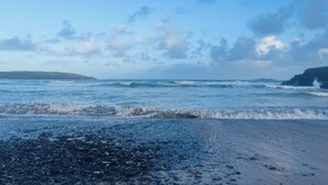 Una playa con olas, piedras y una costa rocosa distante bajo un cielo parcialmente nublado.