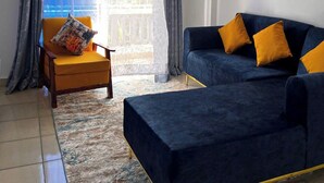 Sala de estar com poltrona amarela, sofá azul com almofadas amarelas e vista para um prédio azul pela janela.