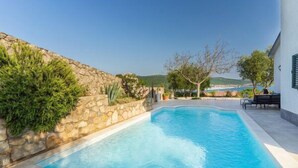 A swimming pool with a stone wall and greenery on one side, a patio area with a table and chairs, and a view of the sea and hills in the background.