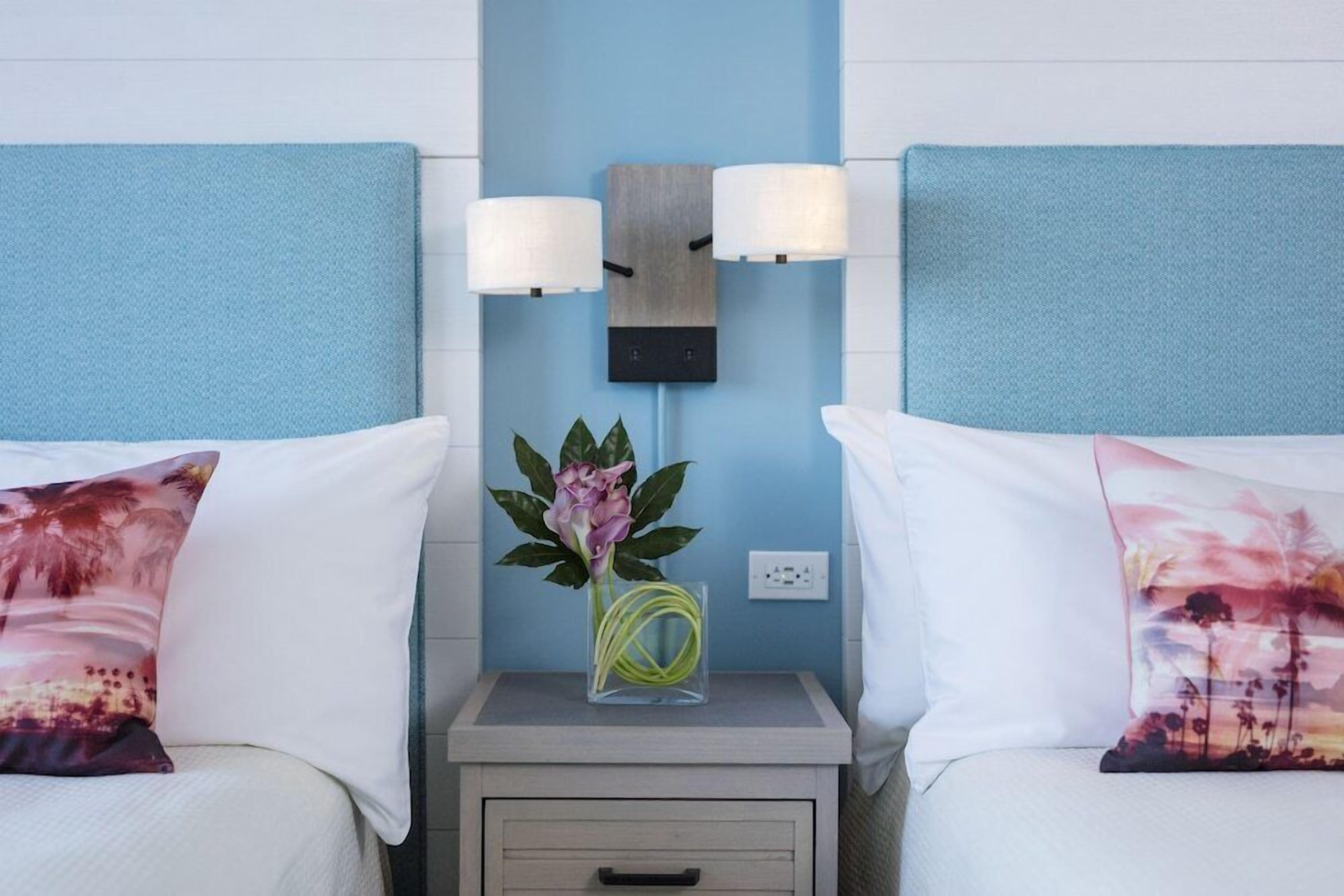 Ein Hotelzimmer mit zwei Betten, jedes mit einem weißen Kissen und einem dekorativen Kissen mit Sonnenuntergangsmotiv. Zwischen den Betten befindet sich ein Nachttisch mit Blumen in einer Vase und einer Wandlampe.