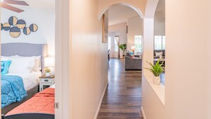 Ein moderner Hotelkorridor mit Holzboden, einem Bett mit blau-orange gestreifter Bettwäsche, einem grauen Sofa und einer Pflanze.