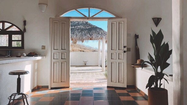 An open doorway leading to a patio with a thatched roof, a potted plant, and a bar area with stools. - Casa Núuk (Todos Santos)