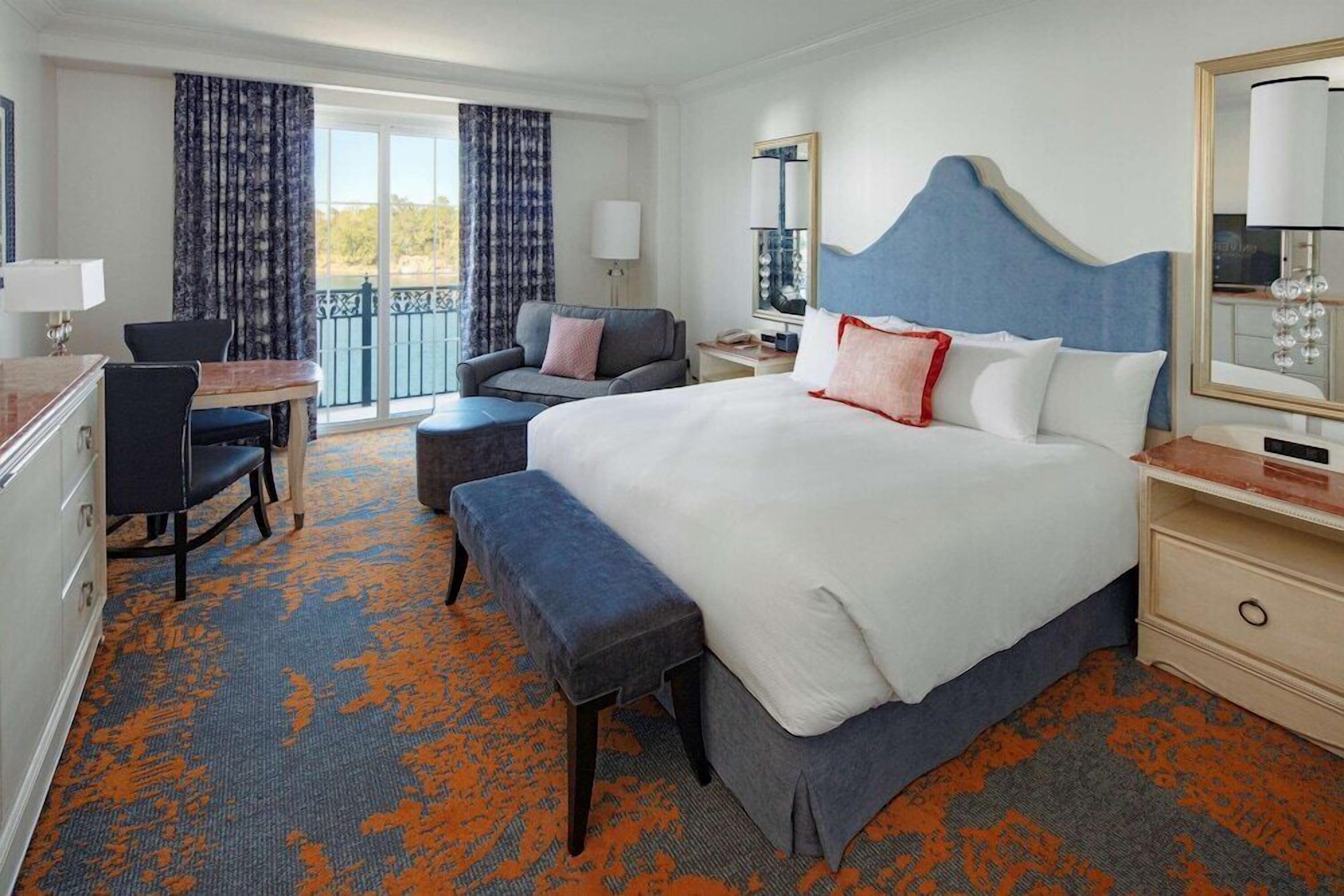 Ein Hotelzimmer mit einem großen Bett, einem Schreibtisch mit Stuhl, einem kleinen Tisch und einem Balkon mit Vorhängen.