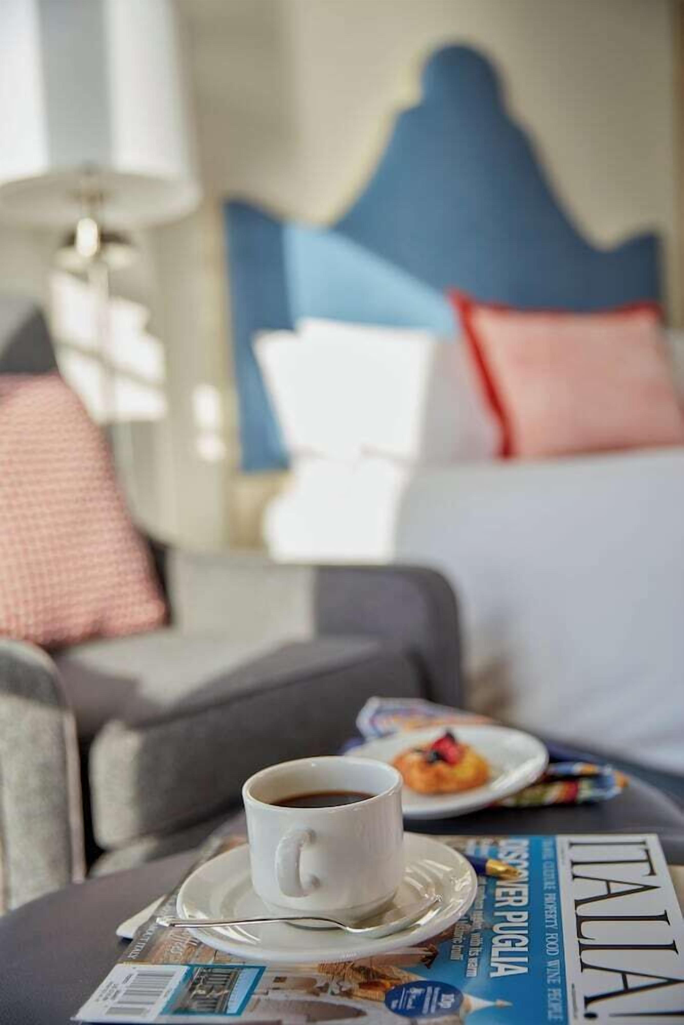 Een hotelkamer met een bed, een bijzettafeltje met een kop koffie en een bord eten, en een tijdschrift op tafel.