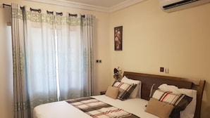 A hotel room with a large bed, a headboard, a nightstand, a wall-mounted air conditioner, and a window with curtains. - Fokuzzy Lodge (Madina)