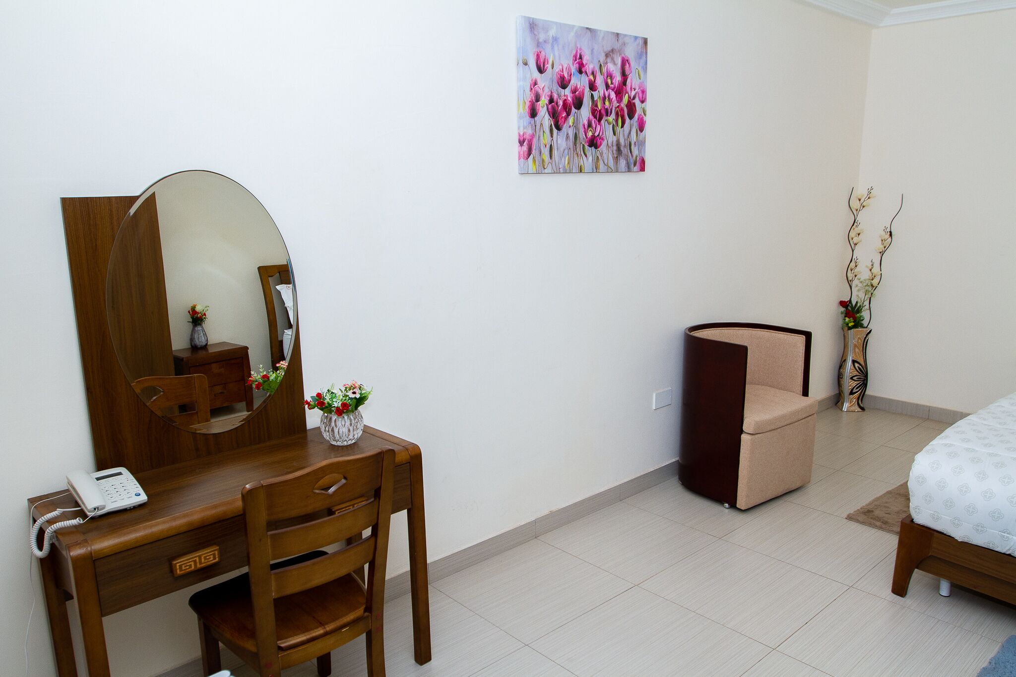 Une chambre d’hôtel avec un bureau en bois, une chaise, un lit, une table de chevet, un miroir, un vase avec des fleurs et un tableau au mur.