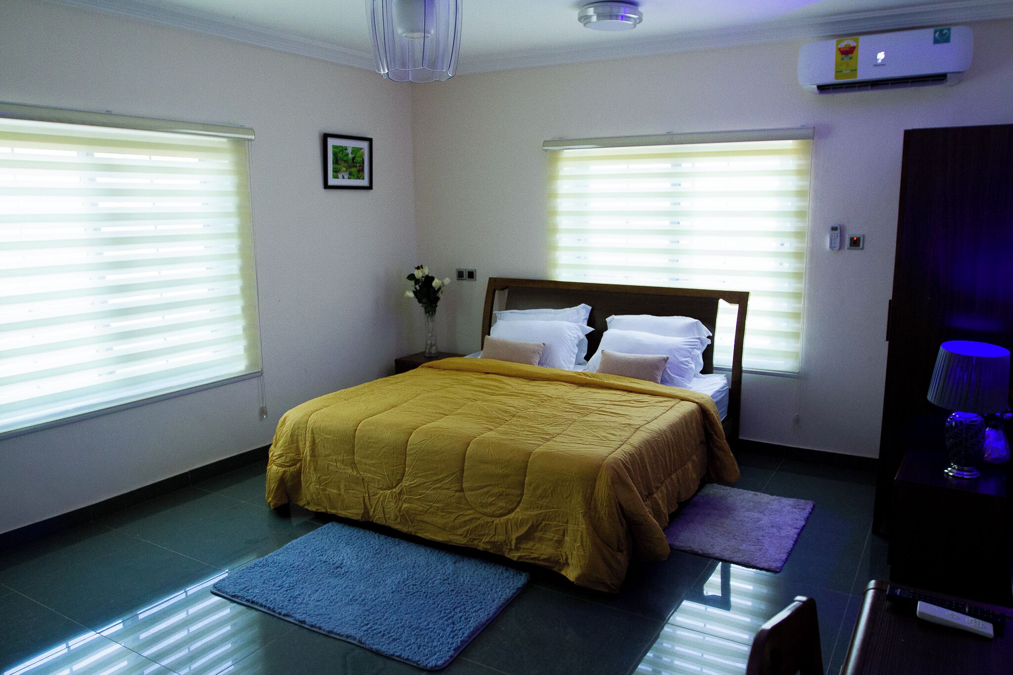 A bedroom with a bed, two windows, a nightstand, a lamp, and a wall-mounted air conditioner.